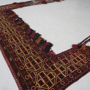 May include: A geometric patterned rug in shades of burgundy, tan, and black. The rug has a rectangular shape with an L-shaped design, featuring intricate patterns and decorative tassels along the edges. The rug is likely handmade.