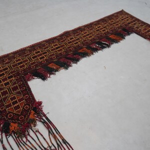 May include: An intricately woven, L-shaped textile with a rich, earthy color palette. The design features geometric patterns in shades of burgundy, gold, and black. The edges are adorned with tassels in matching colors, adding texture and visual interest.