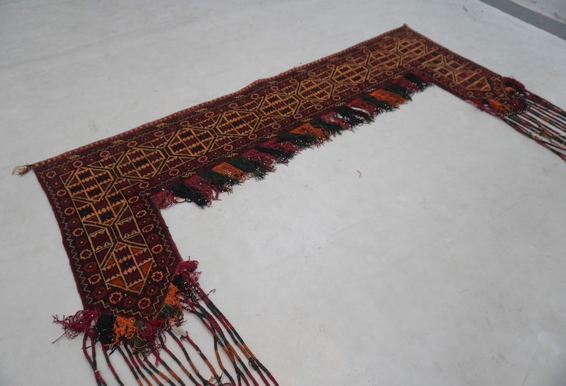 May include: An intricately woven, L-shaped textile with a rich, earthy color palette. The design features geometric patterns in shades of burgundy, gold, and black. The edges are adorned with tassels in matching colors, adding texture and visual interest.
