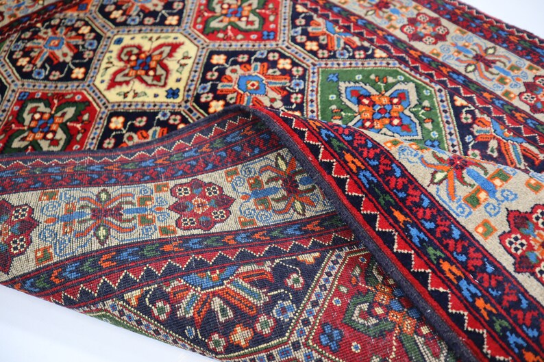 May include: A traditional Persian rug with a colourful geometric pattern. The rug is folded in half, revealing the intricate details of the design. The colours include red, blue, green, and cream.
