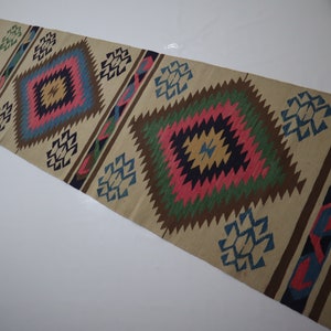 May include: A beige runner rug with a geometric pattern. The design features repeating diamond shapes in pink, blue, green, and brown. The rug has a fringed edge and is suitable for a hallway or entryway.