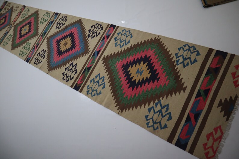 May include: A beige runner rug with a geometric pattern. The design features repeating diamond shapes in pink, blue, green, and brown. The rug has a fringed edge and is suitable for a hallway or entryway.