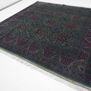 May include: Green and burgundy area rug with an intricate geometric pattern. The rug features a central field with repeating floral motifs and a detailed border. The rug has a fringe on the edges and is placed on a white surface.