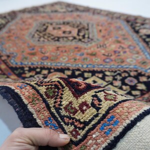 May include: A close-up of a hand holding a small, intricately patterned rug with a central diamond shape. The rug is made of wool and has a fringe on the edges. The colors are predominantly red, blue, and black.