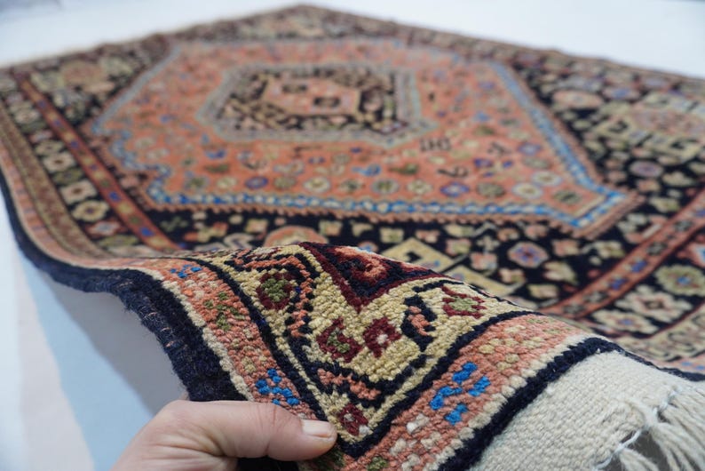 May include: A close-up of a hand holding a small, intricately patterned rug with a central diamond shape. The rug is made of wool and has a fringe on the edges. The colors are predominantly red, blue, and black.