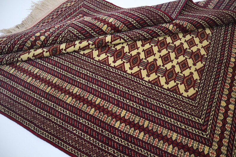 May include: A traditional style rug with a cream background and intricate red, black, and yellow geometric patterns. The rug has a fringe border.