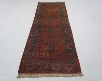 May include: A long, rectangular runner rug featuring a deep red base color with intricate dark blue and black geometric patterns. The rug has a wide border with a repeating scalloped design and includes light-colored fringe at both ends.