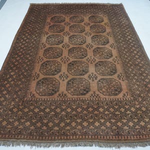 May include: Rectangular brown rug with a central pattern of repeating octagons and floral motifs. The rug has a border with geometric designs and fringe along the edges. The color palette includes shades of brown and tan.