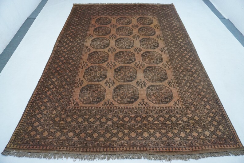 May include: Rectangular brown rug with a central pattern of repeating octagons and floral motifs. The rug has a border with geometric designs and fringe along the edges. The color palette includes shades of brown and tan.