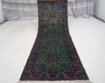 May include: Green runner rug with a detailed pattern. The rug features a central field of green with red and blue accents. The border is a darker shade with intricate designs. The rug has a fringed edge.