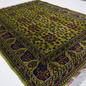May include: A green rug with a traditional Persian design. The rug has a dark blue and red border with a repeating pattern of flowers and leaves. The center of the rug features a large, intricate design with a geometric pattern.