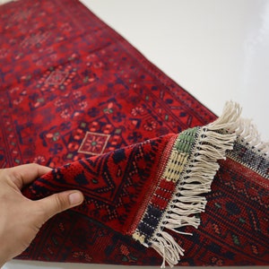 May include: A red runner rug with a traditional Persian design and fringe. The rug is folded in half and a hand is holding the corner.