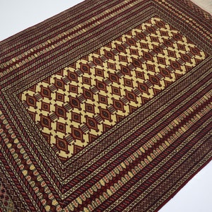 May include: A traditional rug with a red and cream color scheme. The rug features a central diamond pattern surrounded by a border of intricate geometric designs.
