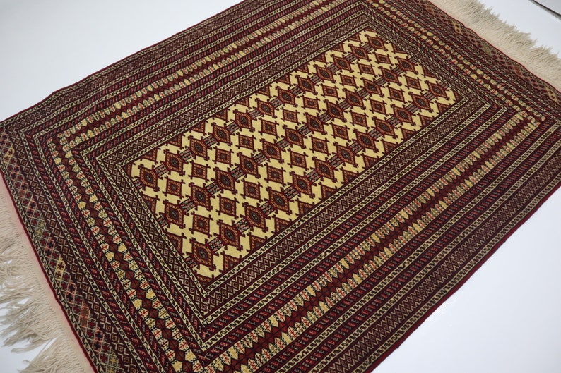 May include: A traditional rug with a red and cream color scheme. The rug features a central diamond pattern surrounded by a border of intricate geometric designs.