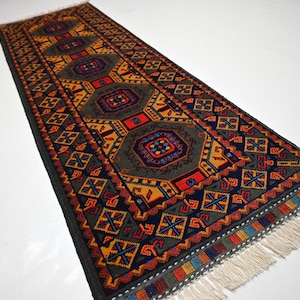 May include: A long, narrow rug with a geometric pattern in shades of red, orange, yellow, blue, and green. The rug has a fringe on both ends.