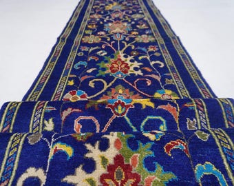May include: A rolled-up, intricately patterned runner rug. The rug features a deep blue background with a central floral design in red, yellow, and green. The edges have a detailed border with complementary colors. The rug is made of a soft material.