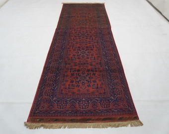 May include: A long, rectangular runner rug featuring a deep red background with intricate navy blue geometric patterns and floral motifs. The rug is framed by a wide navy blue border and finished with short, light-coloured fringe at both ends.