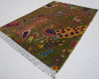 May include: A rectangular green rug with a tree and bird design. The rug features a brown tree trunk with colorful birds, flowers, and a peacock. The rug has a white fringe on the edges.