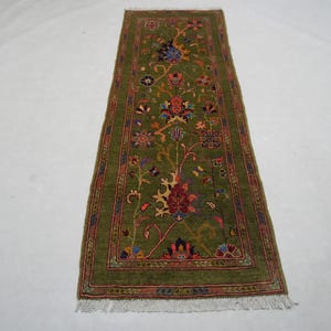 Puede incluir: Una alfombra de pasillo rectangular verde oliva con un estampado floral. La alfombra presenta un diseño central de flores estilizadas en tonos rojos, azules y amarillos. La alfombra tiene flecos en ambos extremos.