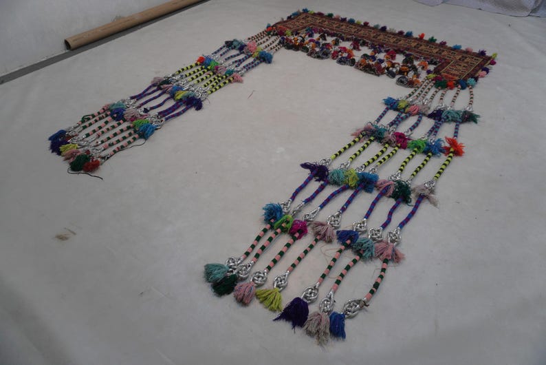 Op de afbeelding: Een sierlijk, handgemaakt textiel met een rechthoekige bovenkant en meerdere strengen kleurrijke kwasten. Het bovenste gedeelte heeft ingewikkelde borduursels met rode, bruine en andere levendige kleuren. De kwasten zijn veelkleurig.