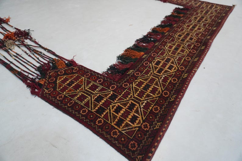 May include: A burgundy and gold geometric patterned rug with tassels. The rug features a repeating diamond and square design in shades of burgundy, gold, and black. The edges are finished with tassels in various colors.