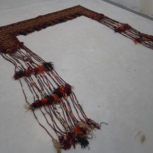 May include: A woven textile with a geometric pattern in shades of brown, red, and orange. Long strands with tassels in matching colors extend from the textile, creating a decorative fringe. The textile is laid out on a white surface.