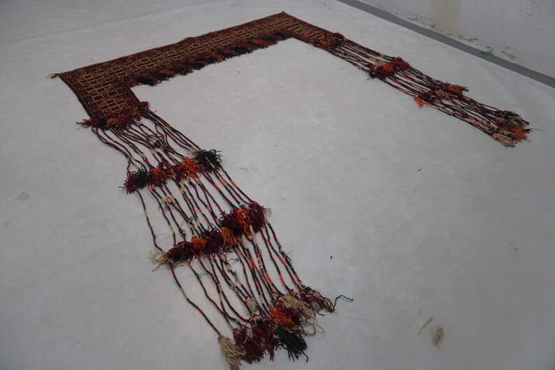 May include: A woven textile with a geometric pattern in shades of brown, red, and orange. Long strands with tassels in matching colors extend from the textile, creating a decorative fringe. The textile is laid out on a white surface.