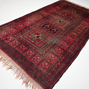 May include: A rectangular, hand-woven rug with a deep red and black geometric pattern. The rug features a central diamond design with smaller repeating motifs and a fringed edge. The rug's dimensions are not visible.