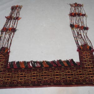 May include: A rectangular, hand-woven rug with a geometric pattern in shades of burgundy, gold, and black. The rug has long, decorative tassels and fringe in various colors. The rug's design is symmetrical and traditional.