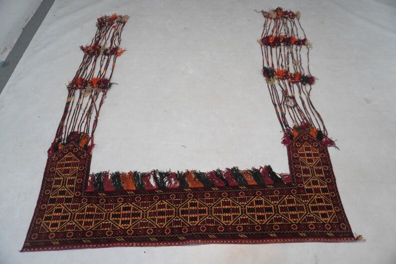May include: A rectangular, hand-woven rug with a geometric pattern in shades of burgundy, gold, and black. The rug has long, decorative tassels and fringe in various colors. The rug's design is symmetrical and traditional.