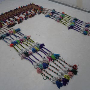 Op de afbeelding: Een sierlijk, rechthoekig textiel met een patroonrand en kleurrijke kwasten. Het textiel heeft een centraal ontwerp met verschillende kleuren en is versierd met meerdere strengen kwasten in tinten blauw, groen, geel en roze.