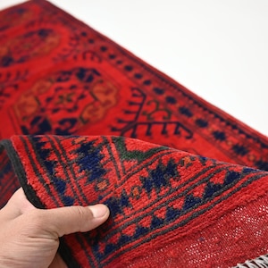 Peut inclure: Un tapis rouge et bleu tissé à la main avec des motifs complexes. Le tapis est plié en deux et a une frange à l'extrémité.