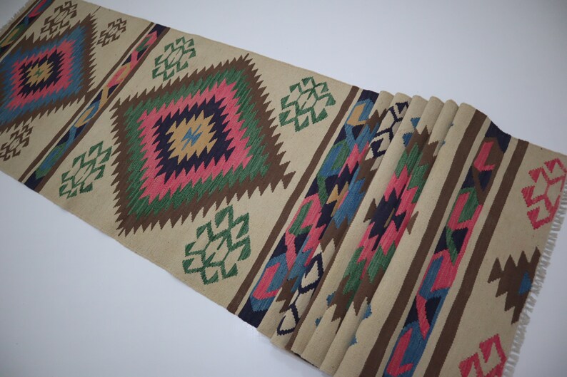 May include: Beige runner rug with geometric patterns in pink, green, blue, and brown. The rug features diamond and zigzag designs, with a fringed edge. Ideal for adding a touch of color and texture to any space.