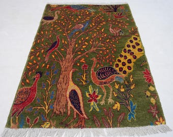 May include: A rectangular green rug with a tree and bird design. The tree is brown with orange leaves, and various birds in red, blue, and yellow are scattered throughout. The rug has a fringe on the bottom edge.