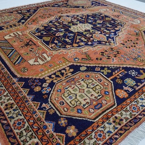 May include: A large rectangular area rug featuring a deep navy blue central medallion and border, contrasted by a salmon-orange field. The rug is decorated with intricate geometric patterns, floral motifs, and stylized animal figures, finished with white fringe.