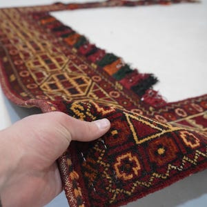 May include: A hand holds a section of a patterned rug. The rug features a geometric design in shades of red, brown, and gold. The edges are finished with tassels in various colors. The rug appears to be made of woven material.