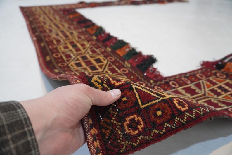 May include: A hand holds a section of a patterned rug. The rug features a geometric design in shades of red, brown, and gold. The edges are finished with tassels in various colors. The rug appears to be made of woven material.