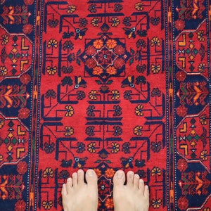 May include: A red rug with a blue and white geometric pattern. The rug has a traditional design with intricate details. Two bare feet are standing on the rug.