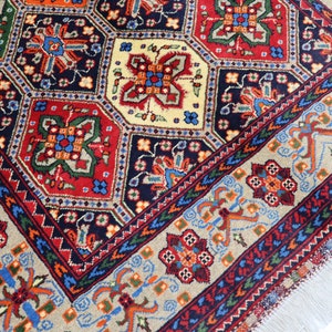 May include: A traditional Persian rug featuring a detailed geometric pattern in shades of red, blue, green, and cream. The rug has a fringed border and is woven with intricate details.