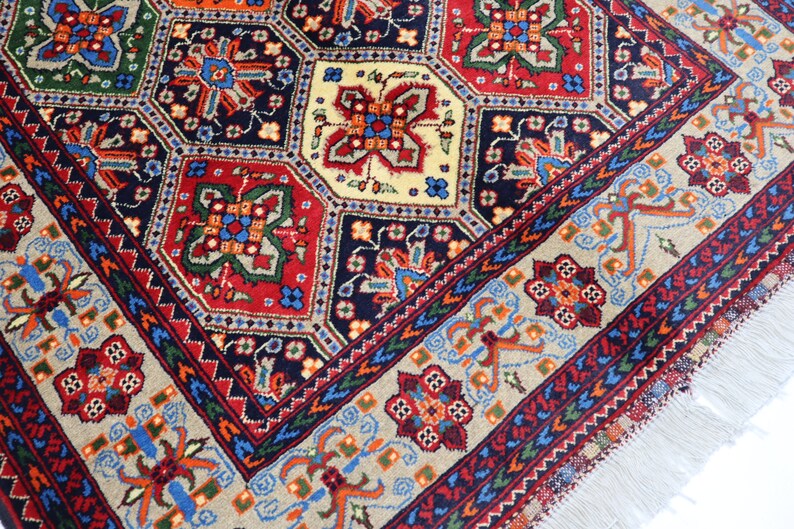 May include: A traditional Persian rug featuring a detailed geometric pattern in shades of red, blue, green, and cream. The rug has a fringed border and is woven with intricate details.