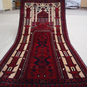 Puede incluir: Una alfombra rectangular roja y beige con un diseño central que presenta una estructura similar a una mezquita y motivos de pájaros estilizados. La alfombra tiene un borde detallado y probablemente está hecha de material tejido.