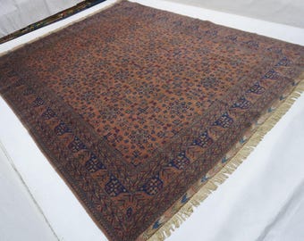 May include: A large rectangular area rug featuring a terracotta orange field with a repeating geometric pattern in dark blue and cream. The rug is framed by a wide dark blue border with stylized floral motifs and finished with cream fringe.