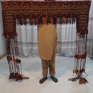 May include: A rectangular, ornate, dark red textile with geometric patterns and long, dangling tassels. The textile is held up, showcasing its intricate design and decorative fringe.