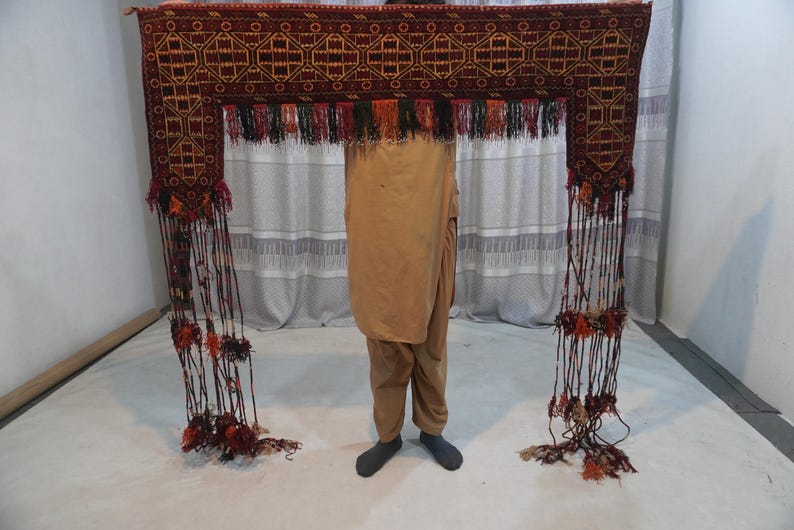 May include: A rectangular, ornate, dark red textile with geometric patterns and long, dangling tassels. The textile is held up, showcasing its intricate design and decorative fringe.