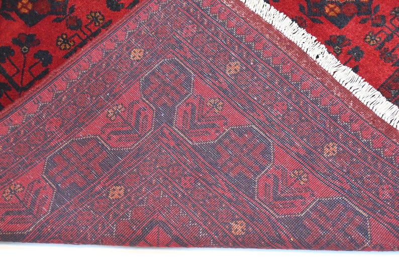 May include: Close-up of the back of a red rug with a dark blue geometric pattern. The rug has a white fringe.