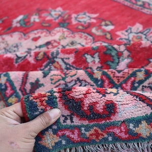 May include: A close-up of a red, pink and blue patterned rug with a hand holding the edge of the rug. The rug has a fringed border.