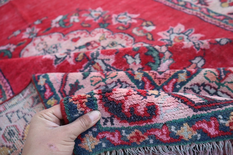 May include: A close-up of a red, pink and blue patterned rug with a hand holding the edge of the rug. The rug has a fringed border.