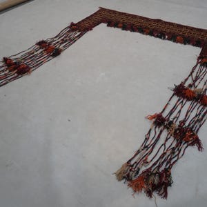 May include: A decorative textile piece with a rectangular woven top and long, fringed tassels. The piece features a brown and red color scheme with accents of orange and cream. The tassels are adorned with small, colorful pom-poms.