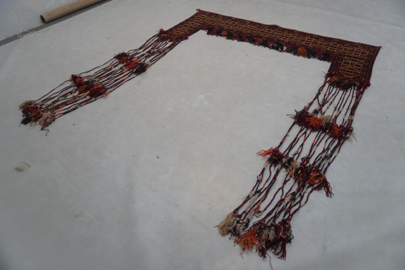 May include: A decorative textile piece with a rectangular woven top and long, fringed tassels. The piece features a brown and red color scheme with accents of orange and cream. The tassels are adorned with small, colorful pom-poms.