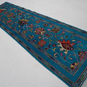 May include: A rectangular, turquoise runner rug with a floral pattern. The rug features a central field of turquoise with a repeating floral design in shades of red, orange, yellow, and brown. The border is a darker turquoise with a repeating pattern.